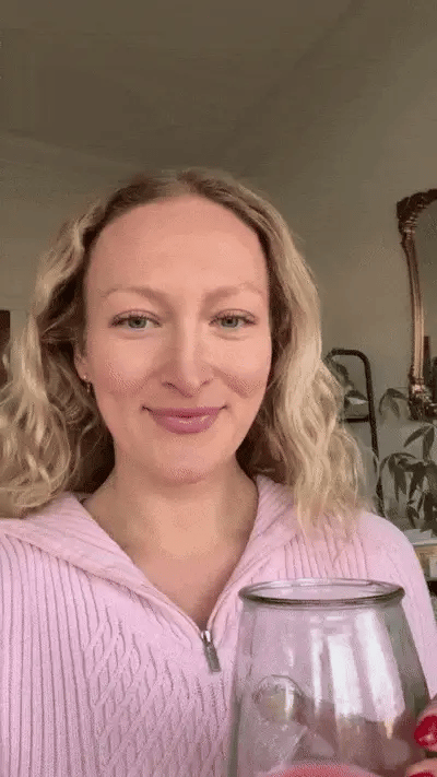 A person with wavy blonde hair smiles while holding a smoothie in a glass. They wear a light pink top, standing in a room with a mirror and plants in the background.