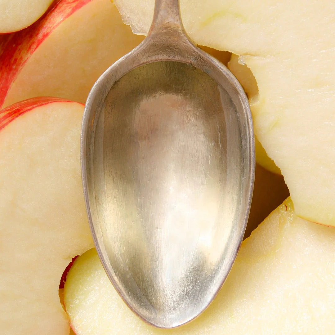 A close-up of a silver spoon resting on slices of fresh, red apple. The spoon reflects light, complementing the apples crisp, juicy texture. The apple slices have a light red skin and are thinly cut.