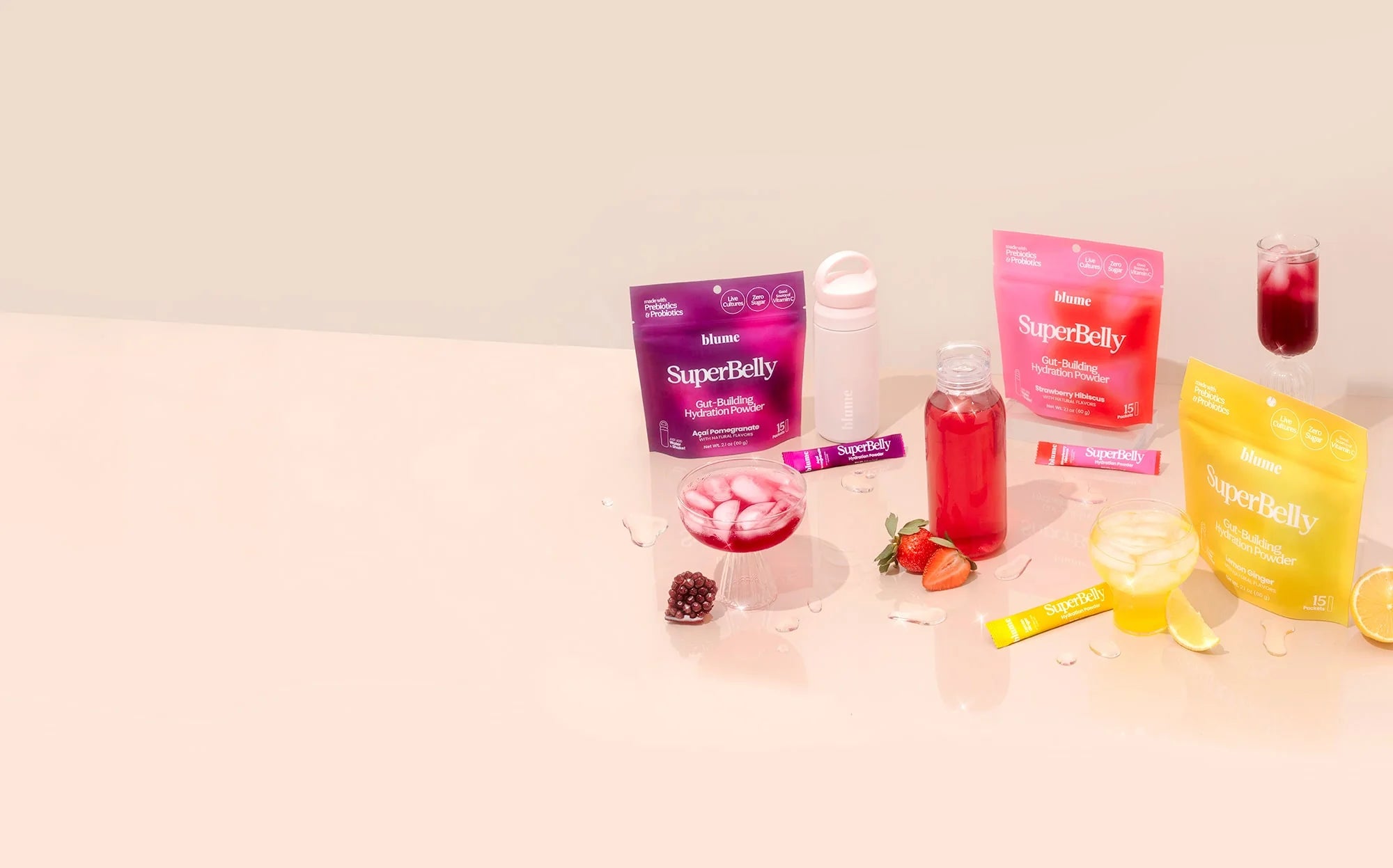 Colorful packets of SuperBelly supplements are arranged on a light surface alongside a glass pitcher filled with red liquid, a cup topped with ice cubes, a water bottle, and scattered pieces of lemon and berries.