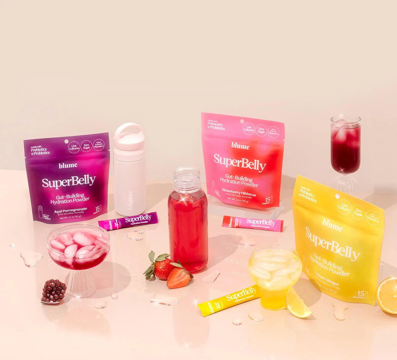 A variety of Blume SuperBelly hydration powder packets, in pink and yellow packaging, are displayed with glasses of red and yellow drinks. Fresh fruit and a water bottle accompany the arrangement on a light surface.