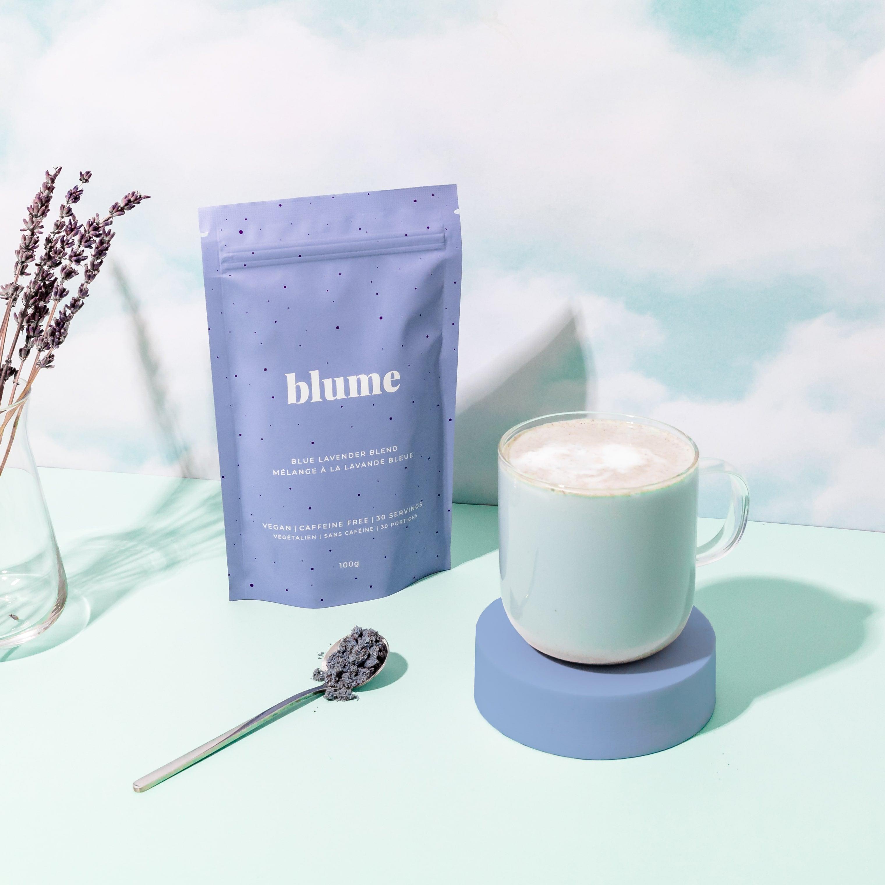 A mug with a frothy blue lavender latte sits on a round coaster beside a Blume Blue Lavender Blend bag. A spoon of loose blend is in front, while decorative calming lavender sprigs enhance the soft blue background, making it an effective anxiety reliever.