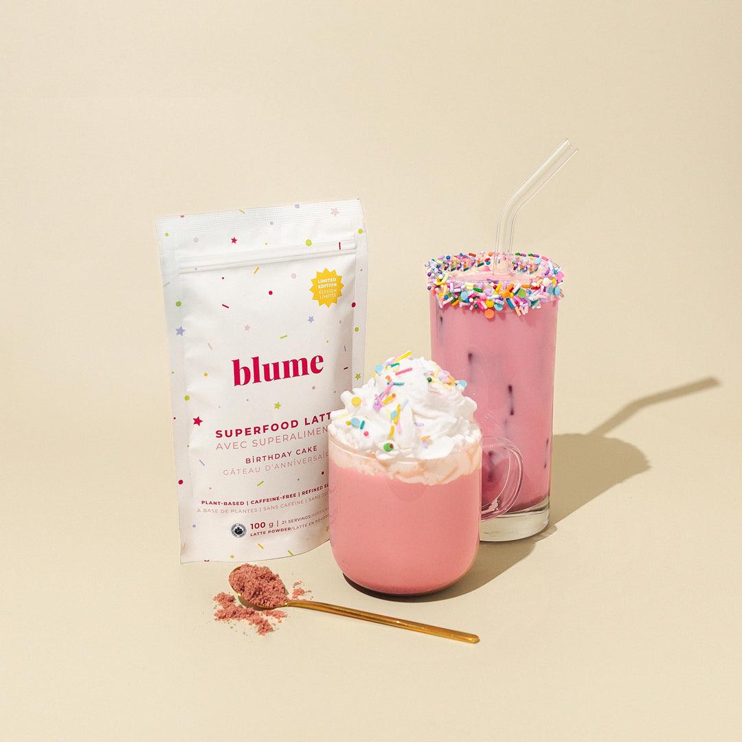 A pouch of Blumes Birthday Cake Latte Blend is next to two pink lattes: one in a mug with whipped cream and another in a glass with rainbow sprinkles and a straw. A spoonful of pink powder from the Organic Ingredients is placed in front.
