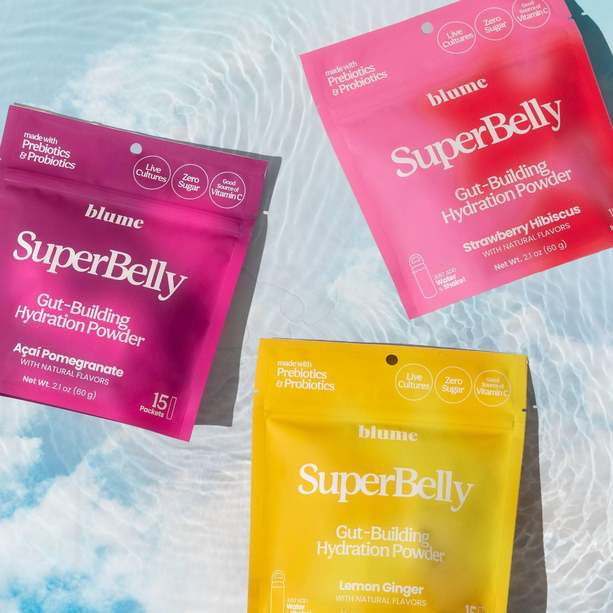 Three SuperBelly gut-building hydration powder packets by Blume are displayed on a light blue, water-like background. The flavors include Acai Pomegranate (pink), Strawberry Hibiscus (red), and Lemon Ginger (yellow). Each pack features nourishing superfoods, potent probiotics, and zero sugar for optimal gut health.