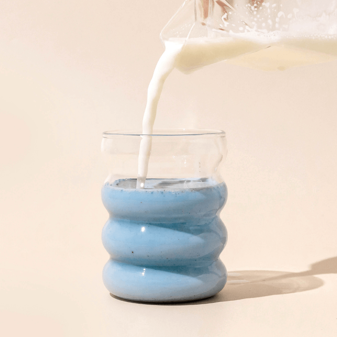 Milk flows into Blumes Bubble Cup, blending with its light blue iced latte against a minimalist beige background, creating a visually captivating contrast.