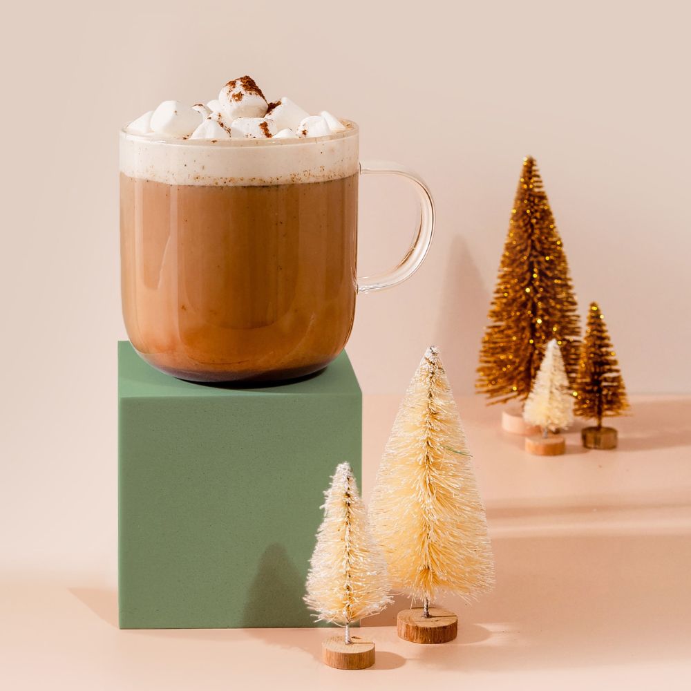 A cup of Blumes Mint Cocoa Blend with whipped cream and marshmallows rests on a green block. Beside it, decorative pine trees in gold and cream adorn a beige surface, creating a cozy, festive ambiance.