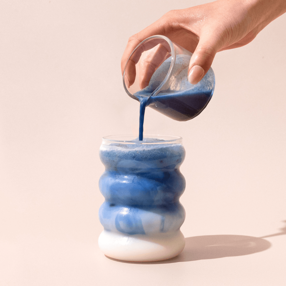 A hand pours a vibrant blue liquid from a beaker into Blumes Bubble Cup, featuring an iced latte with a frothy top, creating mesmerizing swirls against a soft, neutral backdrop that enhances the drink’s vivid colors.