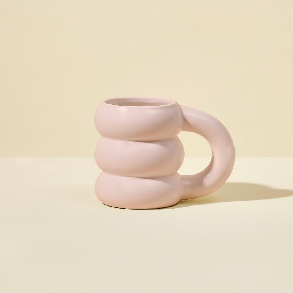 The Cloud Mug by Blume is a limited edition light pink ceramic mug with an oversized circular handle and wavy stacked design. Set against a beige background, this uniquely shaped piece is both functional and fashionable.