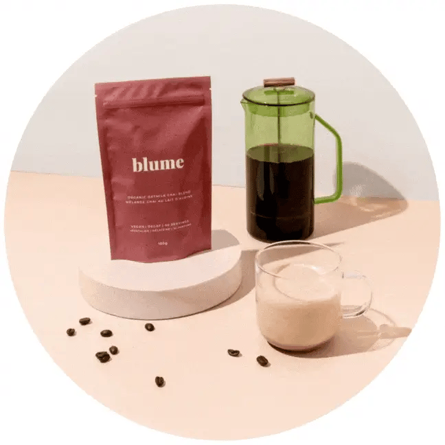 A red bag labeled blume stands on a round white platform next to a glass French press filled with coffee. A clear mug contains a frothy beverage. Scattered coffee beans lie on a light surface.
