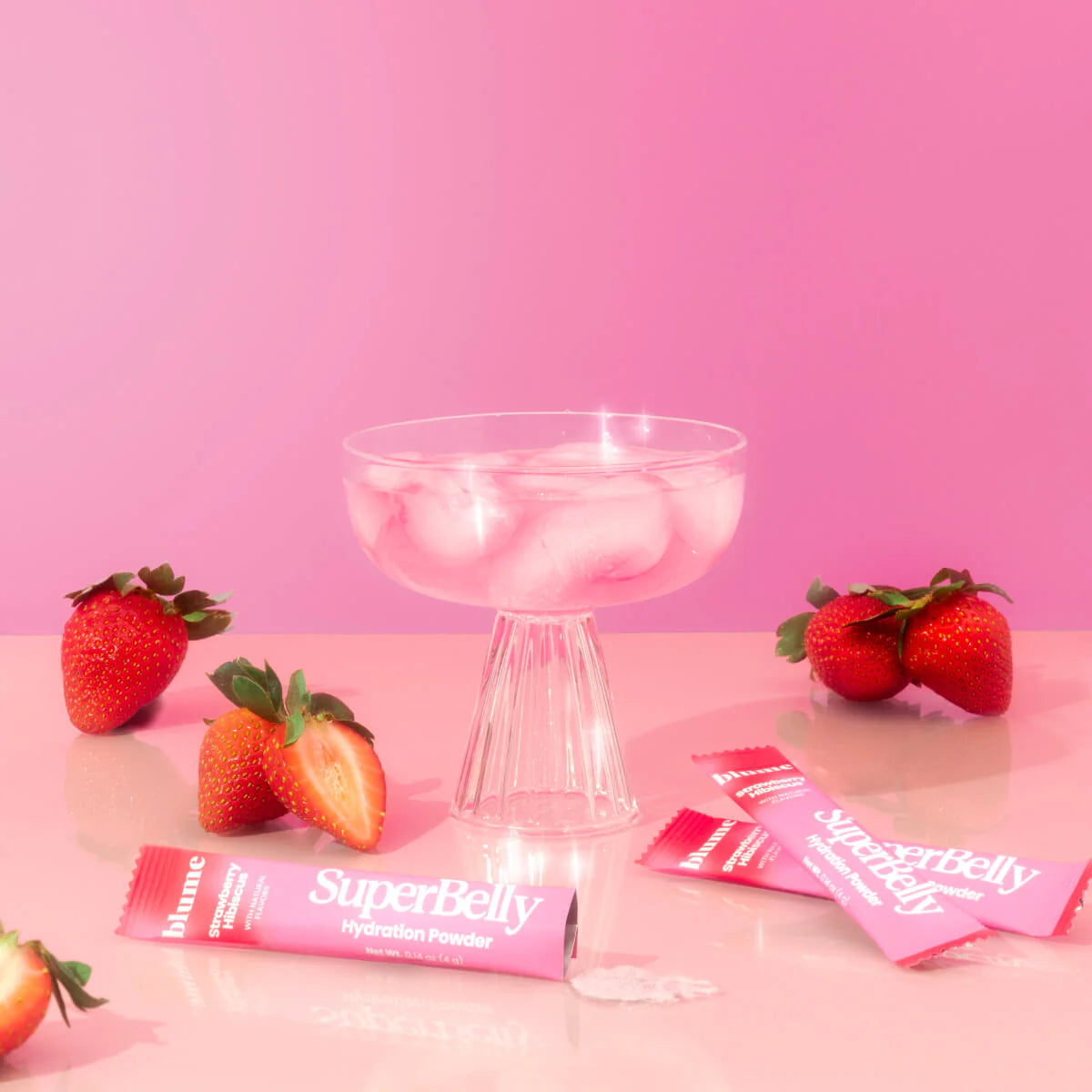 A glass of ice sits on a pink surface surrounded by strawberries and pink sachets labeled SuperBelly Hydration Powder. The background is a solid pink, enhancing the vibrant red of the strawberries.