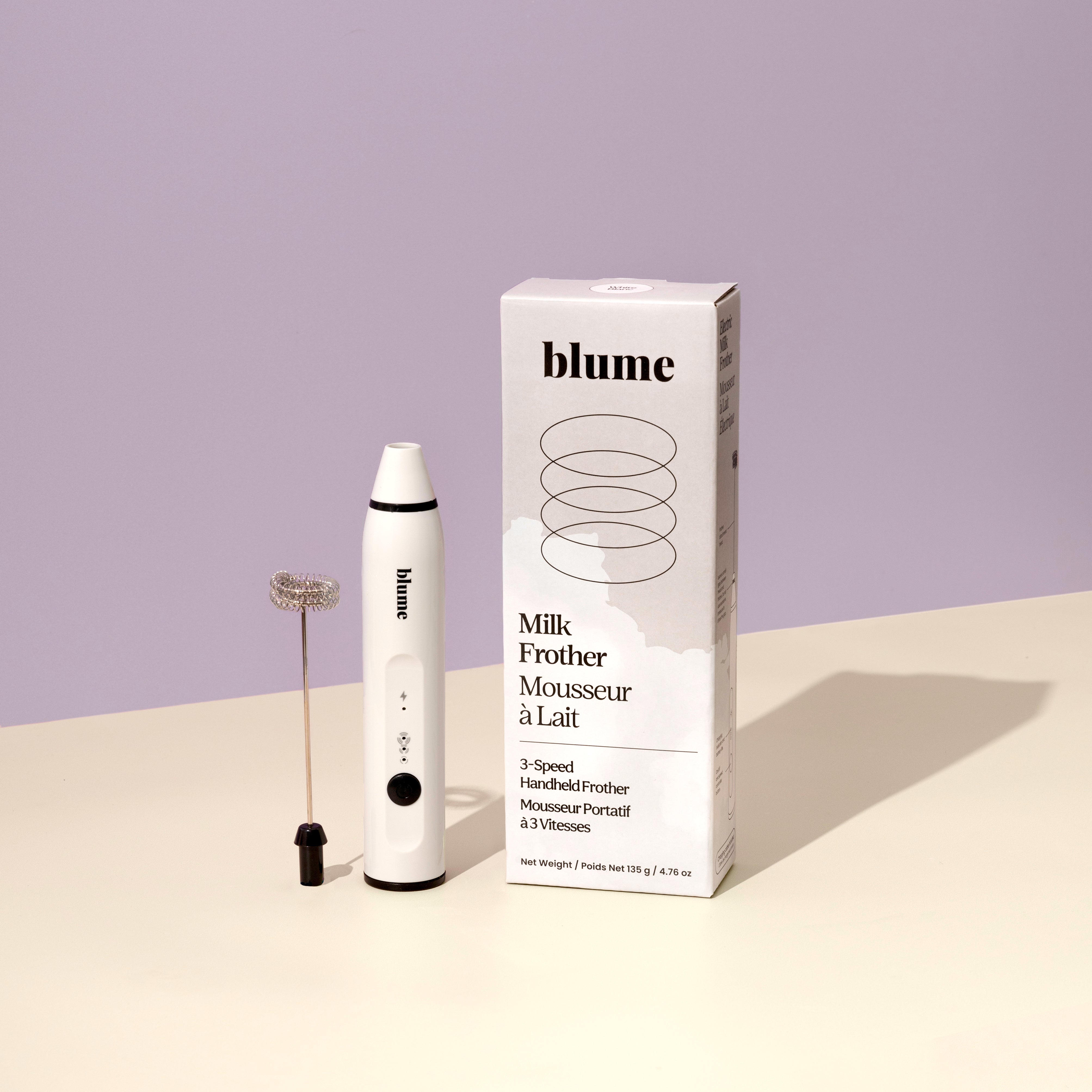 A white handheld milk frother, the Blume 3-Speed Milk Frother, is USB rechargeable and versatile for all frothing needs. It sits beside its packaging on a beige surface with a pale purple background.
