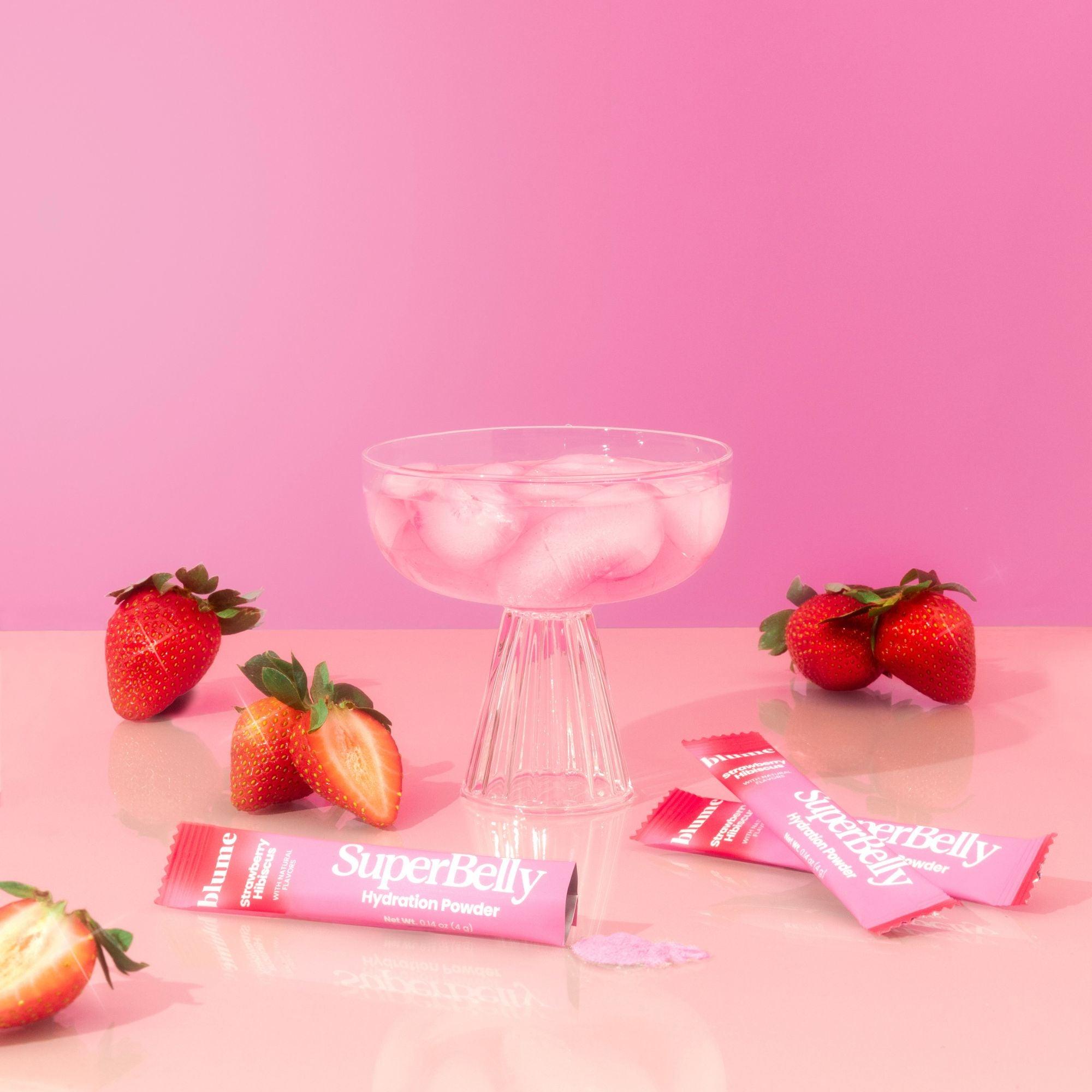Blumes SuperBelly Starter Kit hydration powder sticks, packed with superfoods and probiotics for gut health, are spread on a table with fresh strawberries. A pedestal glass contains ice cubes and pink liquid against a pink gradient background.