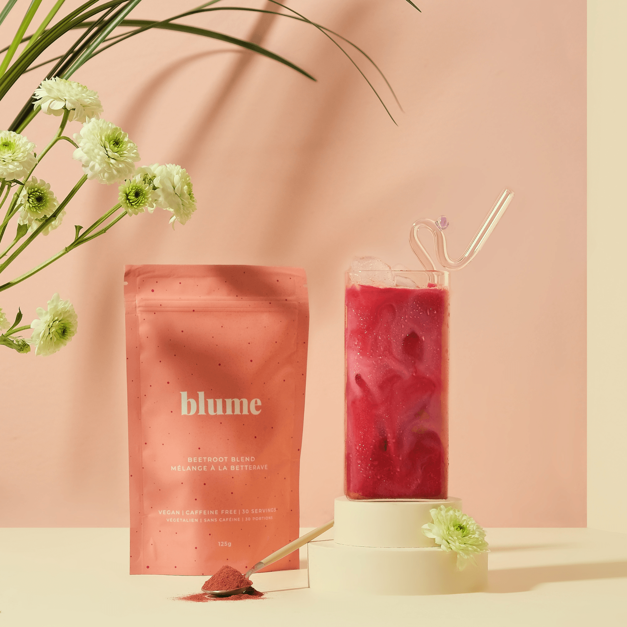 A pink Blume Beetroot Blend package sits beside a tall glass of vibrant pink beetroot latte with ice and a clear straw, all set on a soft pink background, elegantly surrounded by white flowers. This provides a caffeine-free boost.
