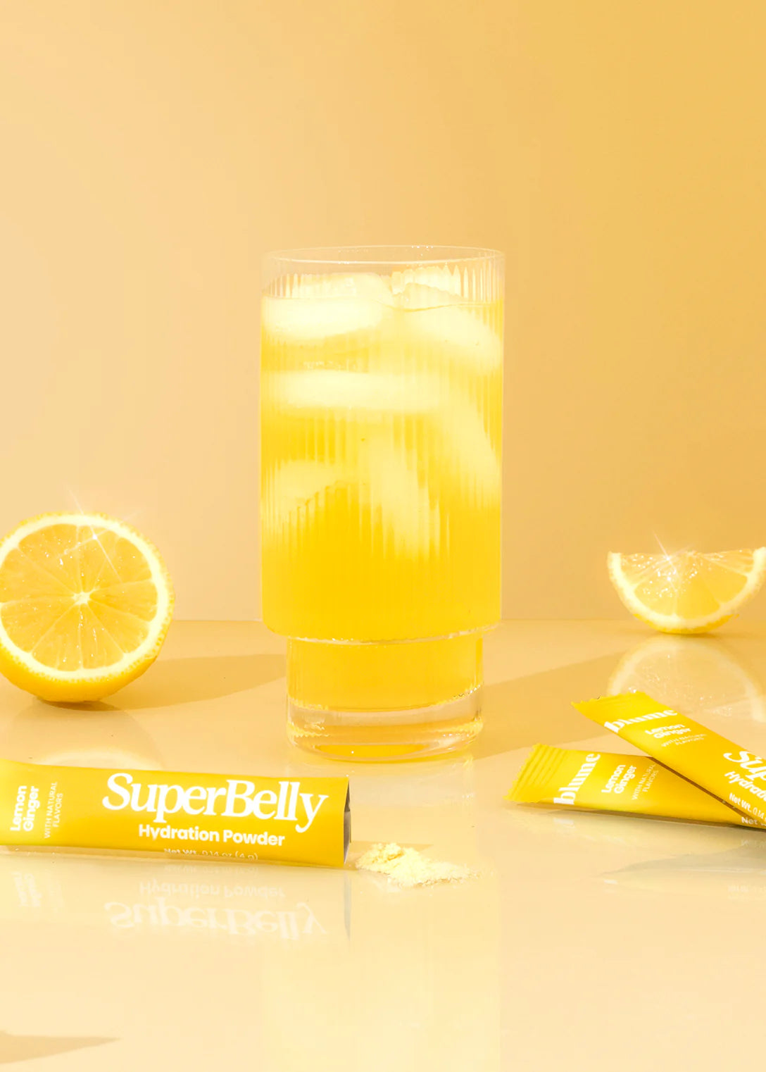A glass filled with iced lemon drink sits on a glossy yellow surface. Next to it are lemon halves and packets labeled SuperBelly Hydration Powder. The overall scene has a bright, fresh, and citrusy aesthetic.