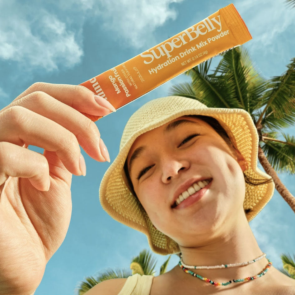 Girl holding a single packet of SuperBelly Mango Passion Fruit Hydration Drink Mix. Cloudy skies and palm trees in the background