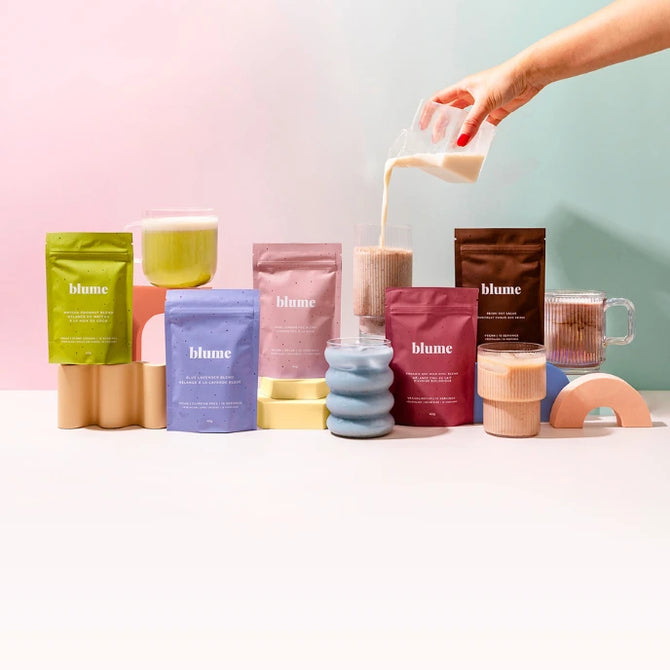 Various colorful packages of Blume drinks are displayed with mugs and glasses filled with beverages. A hand pours milk into a glass. The background features pastel colors in pink and blue.