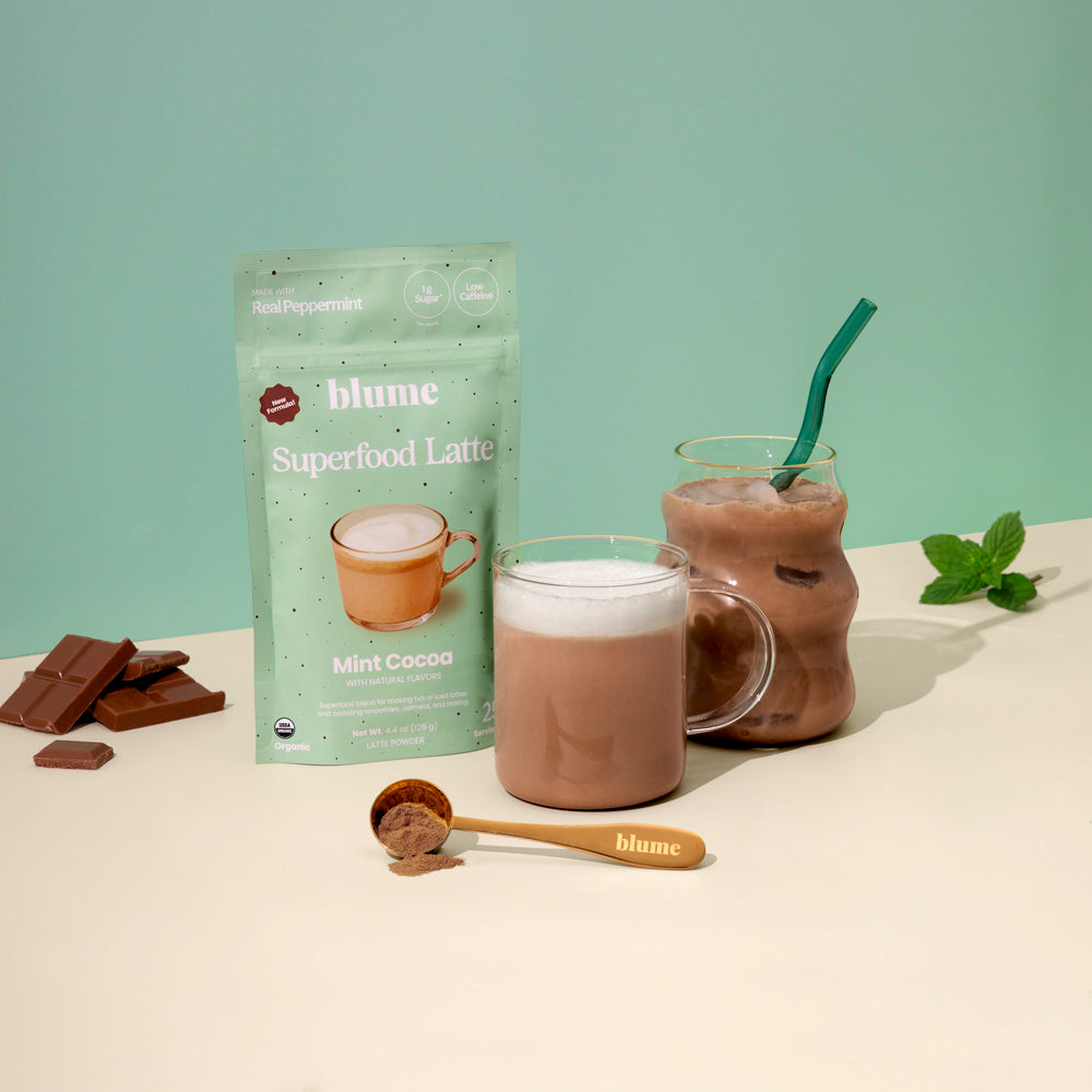 Blumes Mint Cocoa Blend, ideal for dark hot chocolate fans, is in green packaging. Its featured with a glass mug of hot cocoa, a jar of iced peppermint cocoa with a green straw, chocolate pieces, and a wooden scoop of cocoa powder on a light surface against a green background.