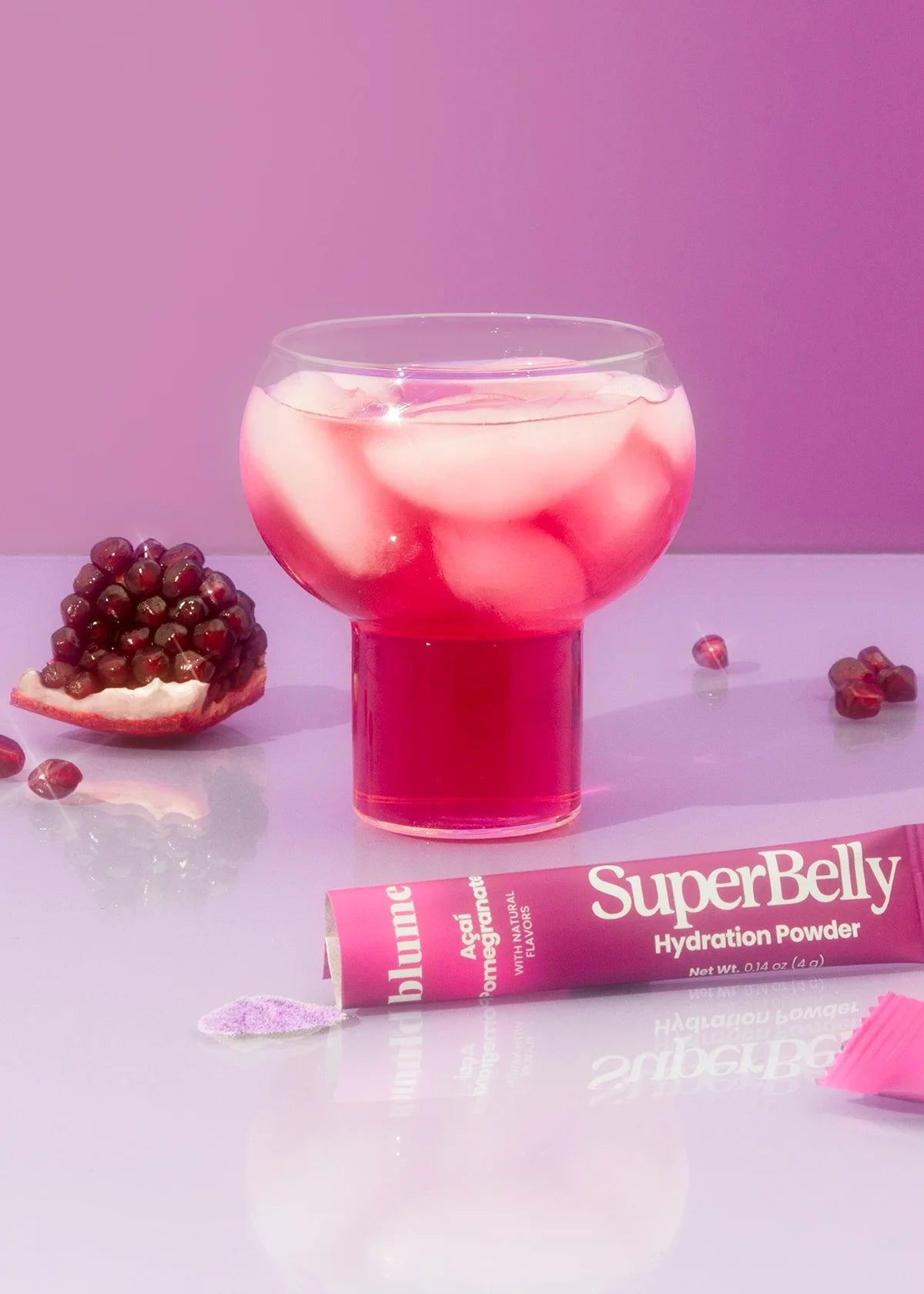 A pink glass filled with a pink iced drink is on a reflective surface. Beside it is a split pomegranate and scattered seeds. A tube labeled SuperBelly Hydration Powder is nearby, with some powder spilled on the surface. The background is pink.