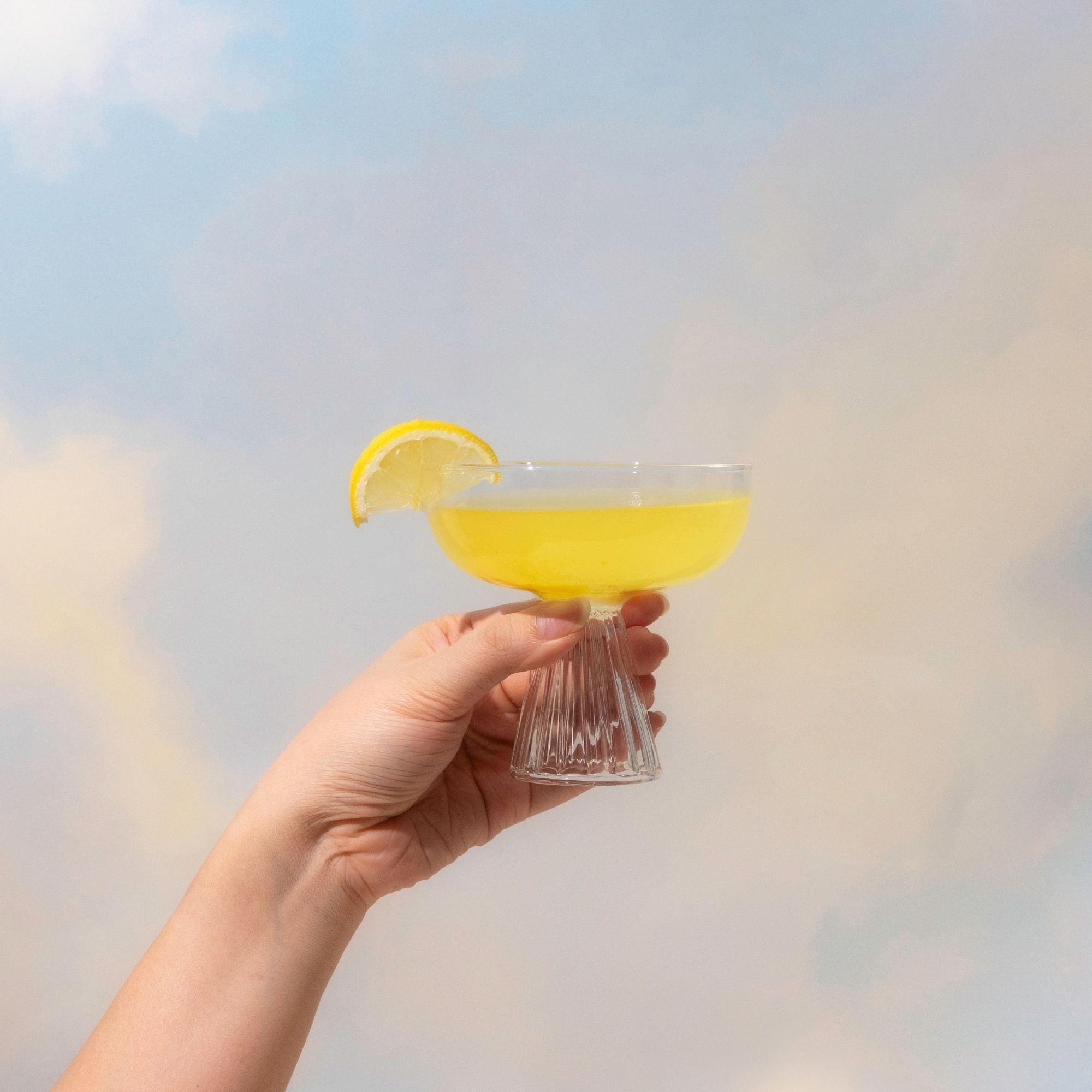 A hand holds a glass of Blumes SuperBelly Lemon Ginger, a probiotic-rich yellow drink, garnished with a lemon slice against a cloudy backdrop.