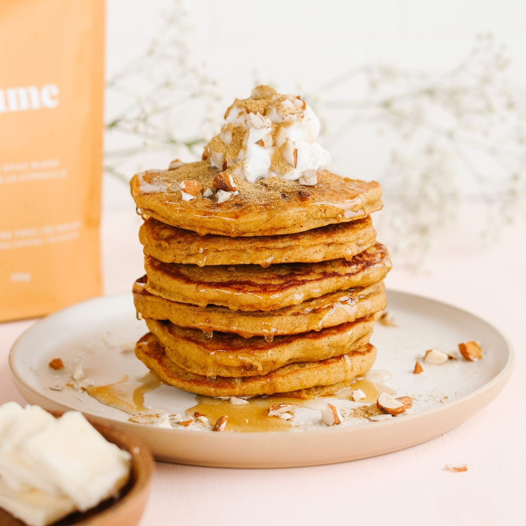 A stack of golden pancakes topped with whipped cream and nuts sits on a beige plate. Honey drips down the sides, and chopped nuts are scattered around. In the background, theres a blurred bag and delicate white flowers.