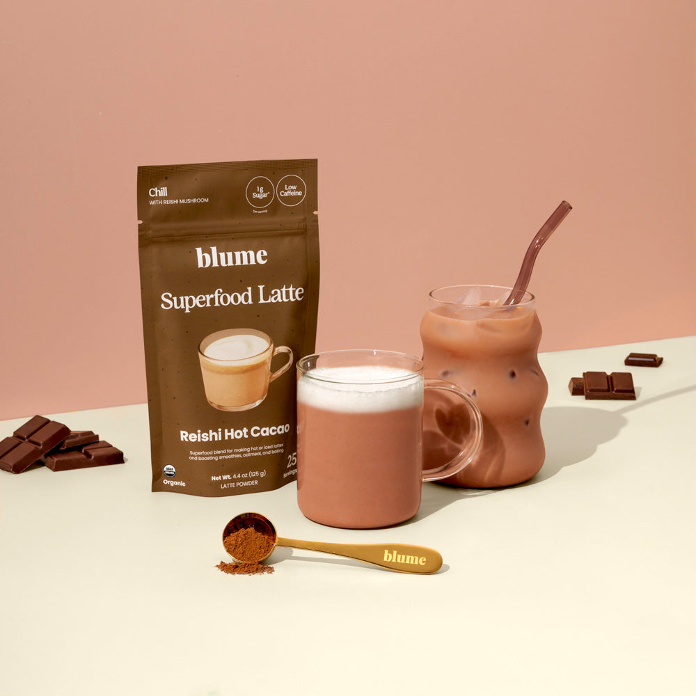 A packet of Blume Superfood Latte mix labeled Reishi Hot Cacao is displayed with a glass mug containing frothed cocoa and an iced chocolate drink with a straw. Chocolate squares and a spoon with cocoa powder complete the arrangement.