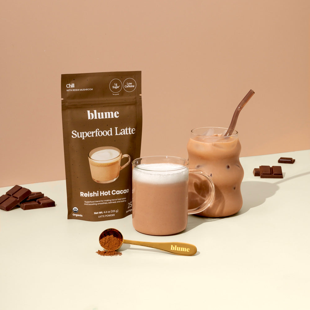 A bag of Blumes Reishi Hot Cacao Blend is featured, showcasing a frothy cacao latte in a clear mug and an iced version in a curvy glass. Next to them, a spoon with adaptogen powder and chocolate pieces rest against a beige background.