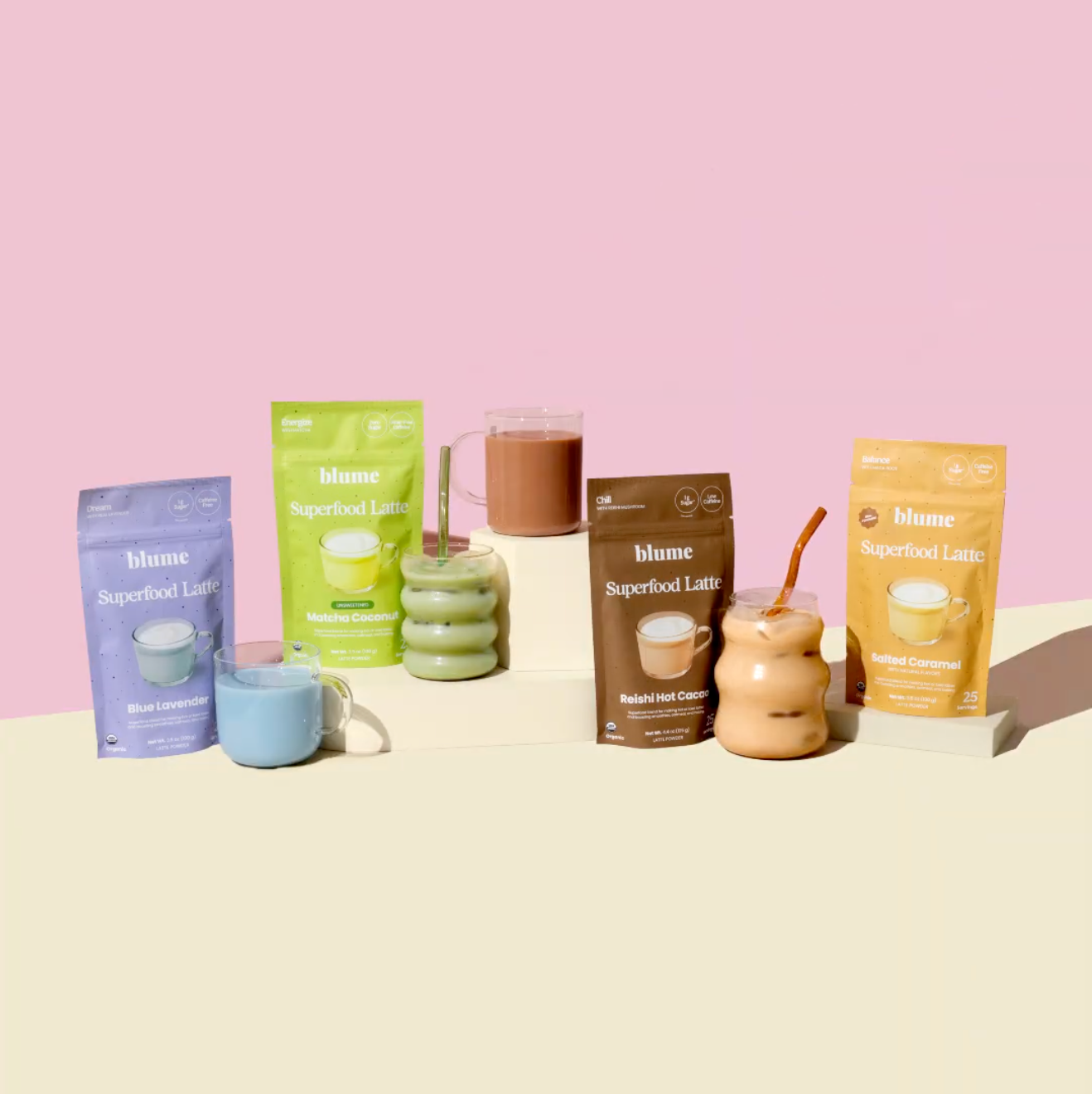 The Blume Latte Essentials Bundle is beautifully arranged on a beige surface, with each colorful pouch of energy-boosting superfoods paired with a mug or glass filled with latte, all against a vibrant pink background.