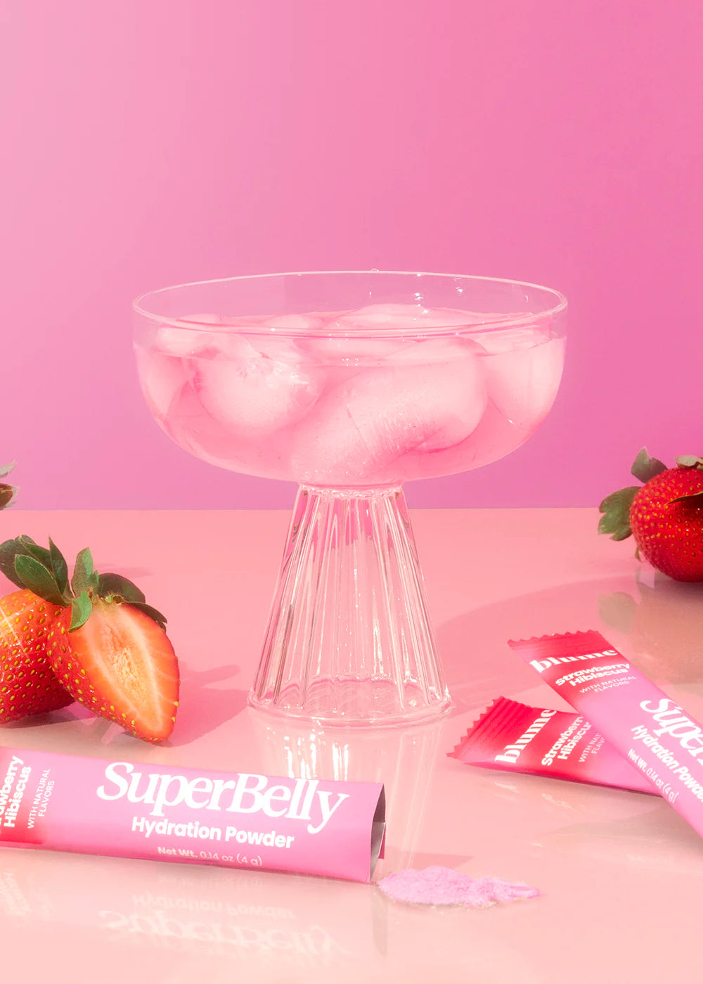 A glass filled with ice cubes and pink liquid sits on a pink surface. Nearby, sliced strawberries and pink packets of SuperBelly Hydration Powder are scattered. The background is a matching pink, creating a vibrant monochromatic look.
