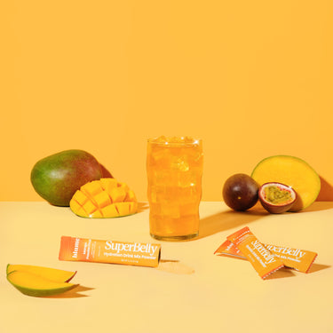 Our newest SuperBelly Gut Health Drink in a glass with ice surrounded by fresh cut mangoes and passionfruit and three packets of SuperBelly Mango Passion Fruit Hydration Drink Mix Powder