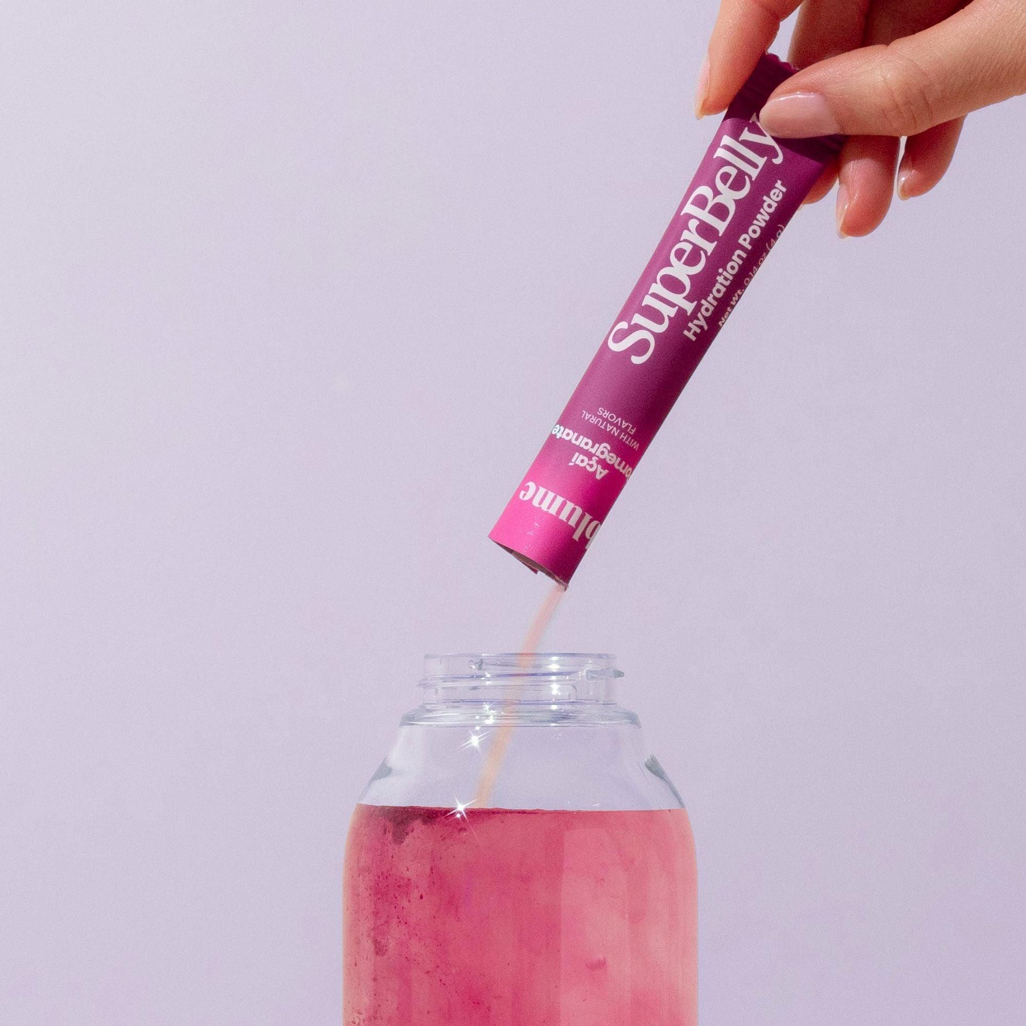 A hand pours a packet labeled SuperBelly Açai Pomegranate by Blume into a glass of pink liquid. This vibrant drink, rich in superfoods and probiotics, enhances gut health against a plain light background, emphasizing the packaging and its colorful contents.