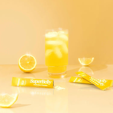 A glass of yellow drink with ice sits amid packets of Blumes SuperBelly Lemon Ginger powder, surrounded by lemon halves and slices on a yellow background, highlighting its fresh citrus flavor and gut-health superfoods.
