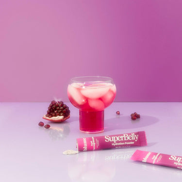A glass of pink beverage with ice sits against a pink backdrop. Beside it are two SuperBelly Açai Pomegranate packets by Blume, offering superfoods for gut health, and a partially opened pomegranate with seeds scattered around.