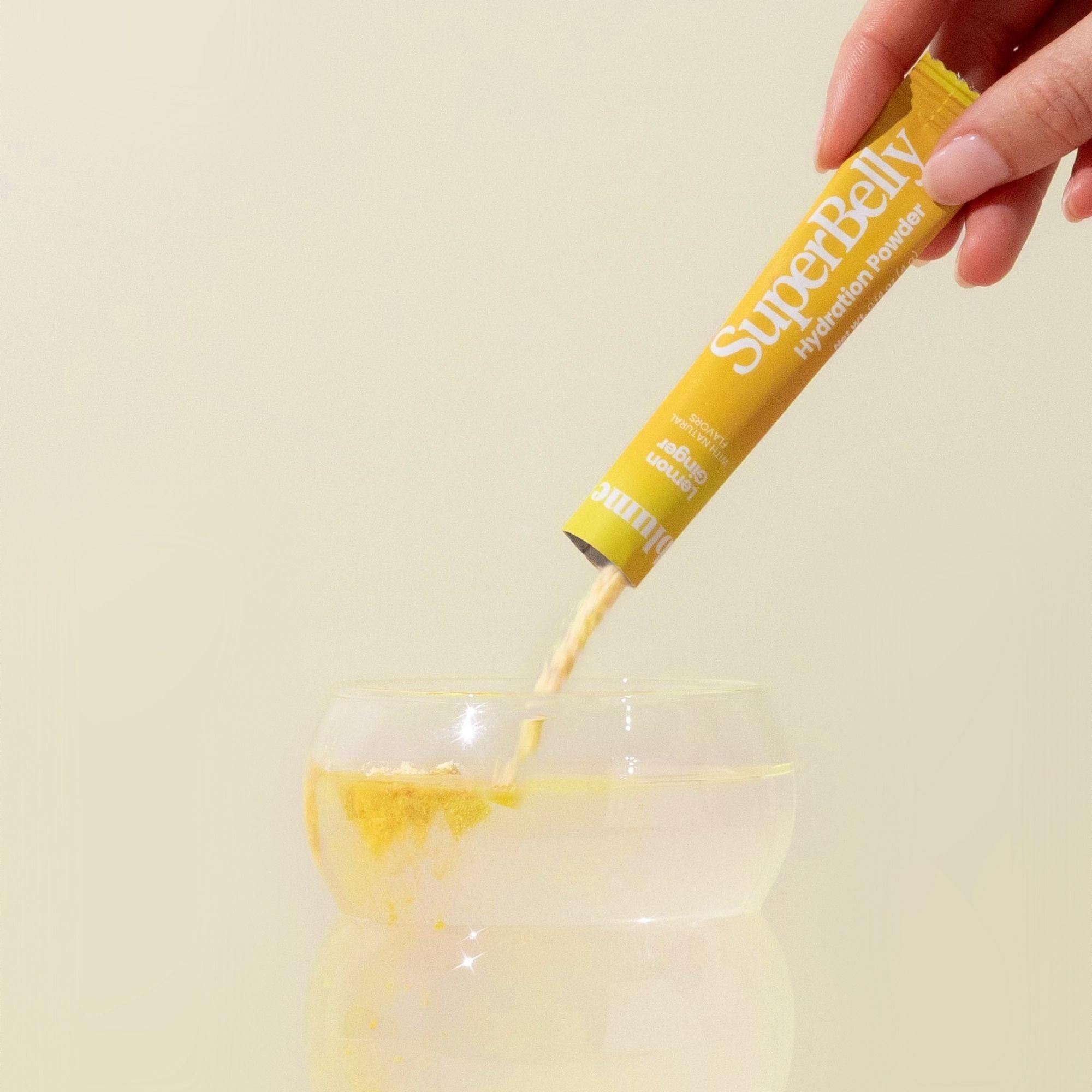 A hand pours a sachet of Blumes SuperBelly Lemon Ginger hydration powder, packed with superfoods and probiotics, into a glass of water against a light off-white background. Mid-pour, the yellow powder forms a pile on the surface, hinting at its gut health benefits.