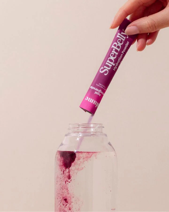 A hand pours a packet of Super Belly hydration powder into a clear bottle of water. The powder creates a vibrant pink swirl as it mixes with the water.