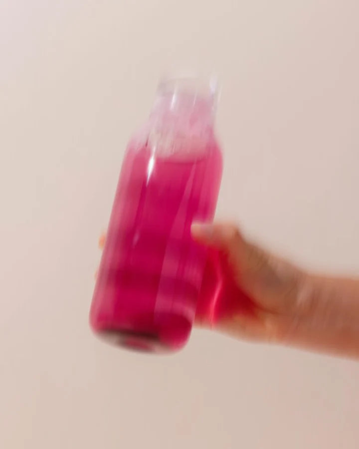 A blurred image of a hand holding a clear bottle filled with bright pink liquid against a pale background.