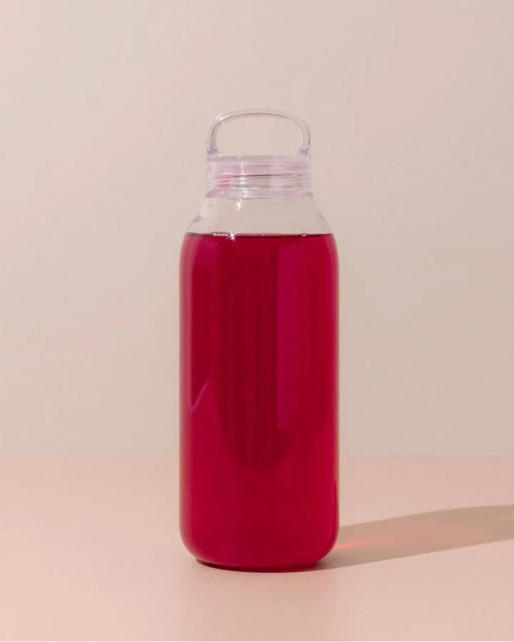 A clear bottle filled with red liquid stands on a beige surface against a light cream background. The bottle has a round, looped handle on its lid. The overall setting has a minimalist and clean appearance.