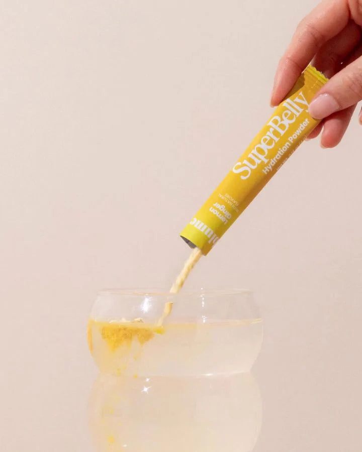 A hand is pouring a yellow hydration powder from a packet labeled SuperBelly into a clear glass filled with water against a neutral background.