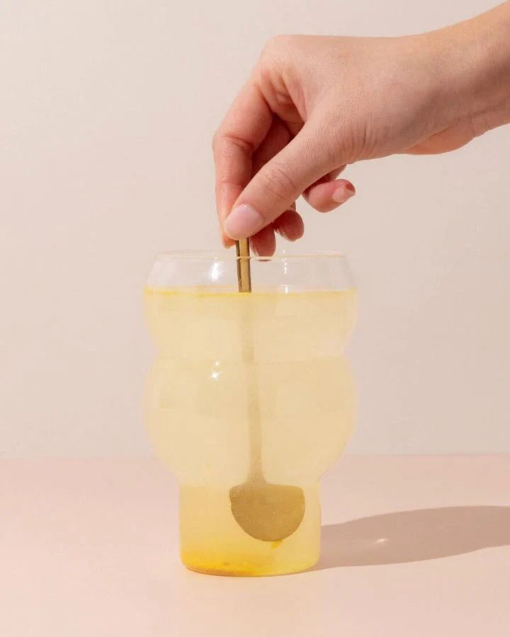 A hand stirring a yellow drink in a uniquely shaped clear glass with a golden spoon. The background is a soft beige color, and the table surface is light pink.