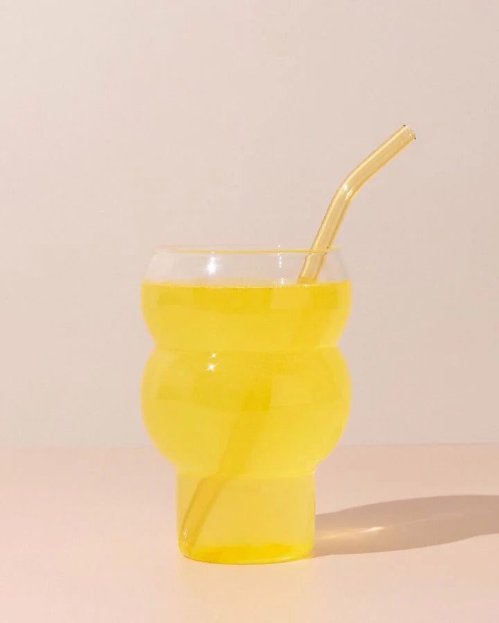 A glass filled with bright yellow liquid features a curvy, artistic shape and a matching bent straw. The glass casts a soft shadow on the light beige surface and background.