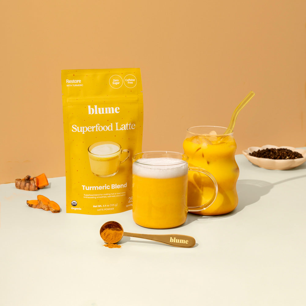 A yellow Blume Turmeric Blend bag sits on a beige backdrop with two glasses of Golden Mylk—one hot and one iced—in front. Nearby are a spoonful of turmeric powder, sliced turmeric, and a bowl of super-spices for added charm.