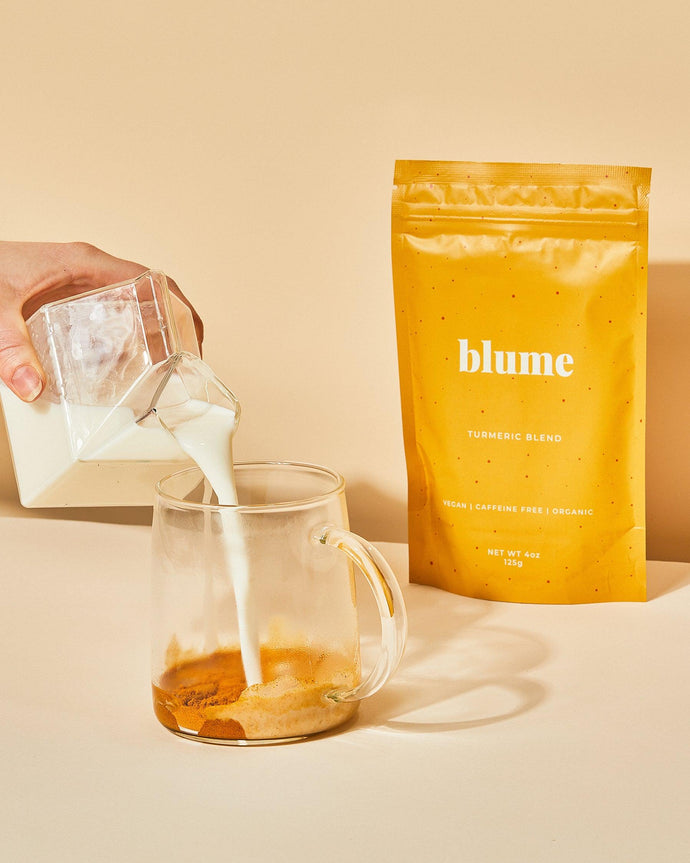 A hand pours a glass of milk into a transparent mug containing a turmeric blend. Beside the mug, there is a yellow package labeled blume Turmeric Blend with text indicating it is vegan, caffeine-free, and organic.