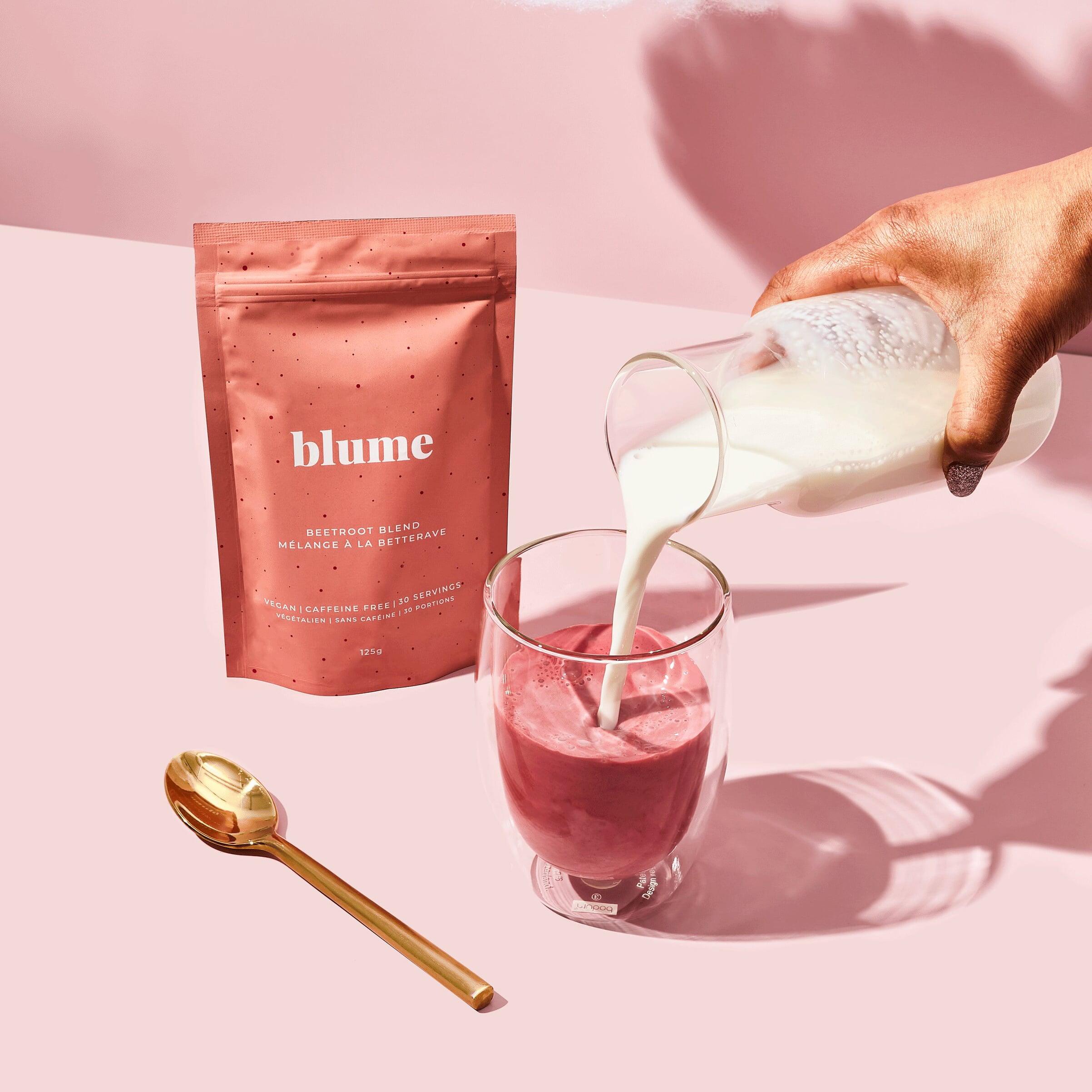 A hand pours milk into a glass of vibrant Beetroot Blend latte, promising a caffeine-free boost. A pink Blume packet is in the background, while a gold spoon on the pink surface hints at its organic ingredients.
