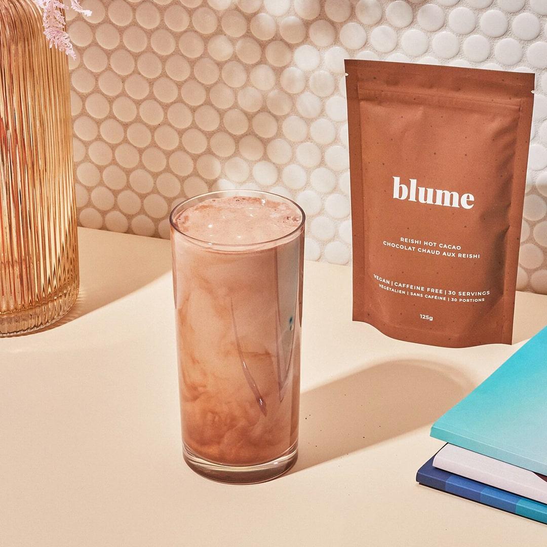A glass of creamy chocolate drink rests on a light table, accompanied by a brown packet labeled Blume Reishi Hot Cacao Blend, an adaptogen with calming properties. The backdrop displays a textured, white circular tile wall, with several books placed to the side.