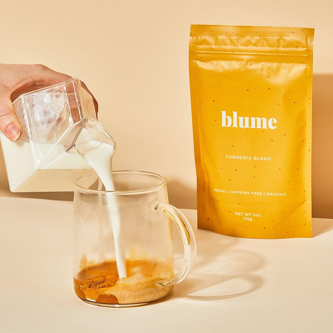 Milk is poured from a glass container into a mug with Blumes Turmeric Blend, creating a soothing turmeric latte. A yellow packet labeled Blume Turmeric Blend sits beside the mug, capturing the essence of Golden Mylk against a beige background.