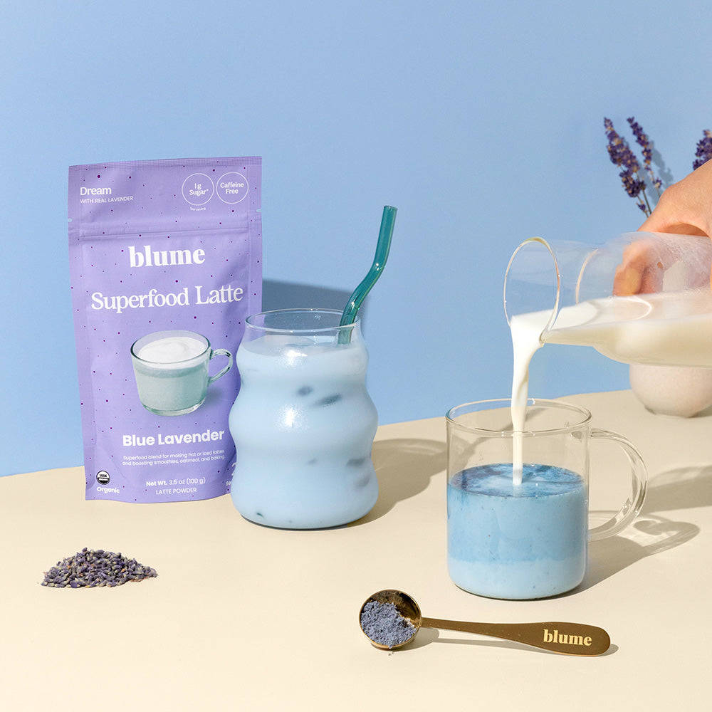 Milk is poured into a glass mug of Blue Lavender latte mix while a wavy glass with iced blue drink glistens nearby. Behind them are sprigs of lavender and a packet labeled Blume The All Day Bundle, evoking tranquility.