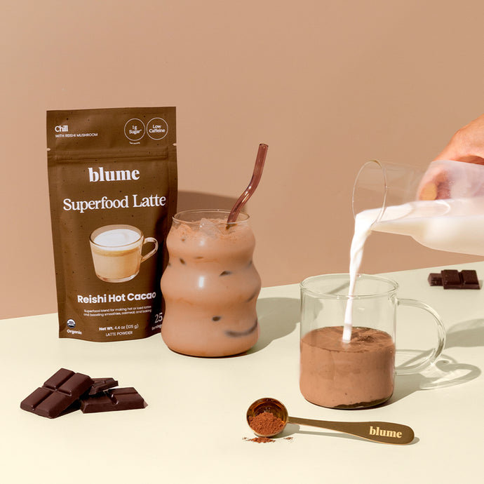 A glass of iced cacao drink and a clear mug of cacao powder being mixed with milk, next to a packet labeled Blume Superfood Latte Reishi Hot Cacao. A gold spoon with cacao powder and chocolate pieces are also visible.
