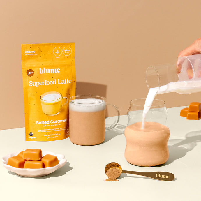 A bag of Blume Superfood Latte Salted Caramel is displayed alongside a glass mug filled with a latte, a jar being filled with milk, a spoon with powder, and caramel cubes on the table.