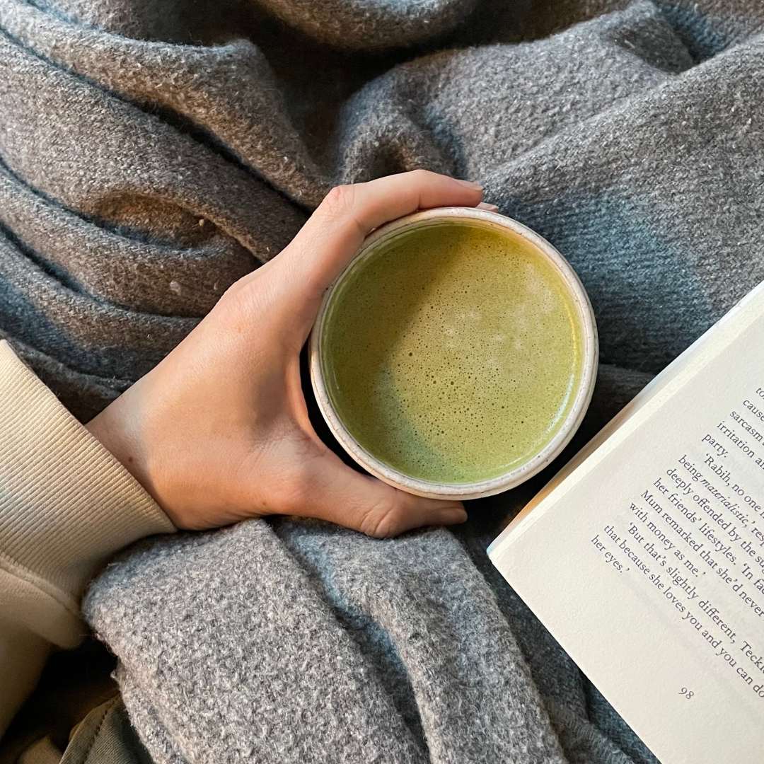 A warm matcha latte made with Blume Matcha Coconut Blend with a book and blanket taken by @brynndolfi