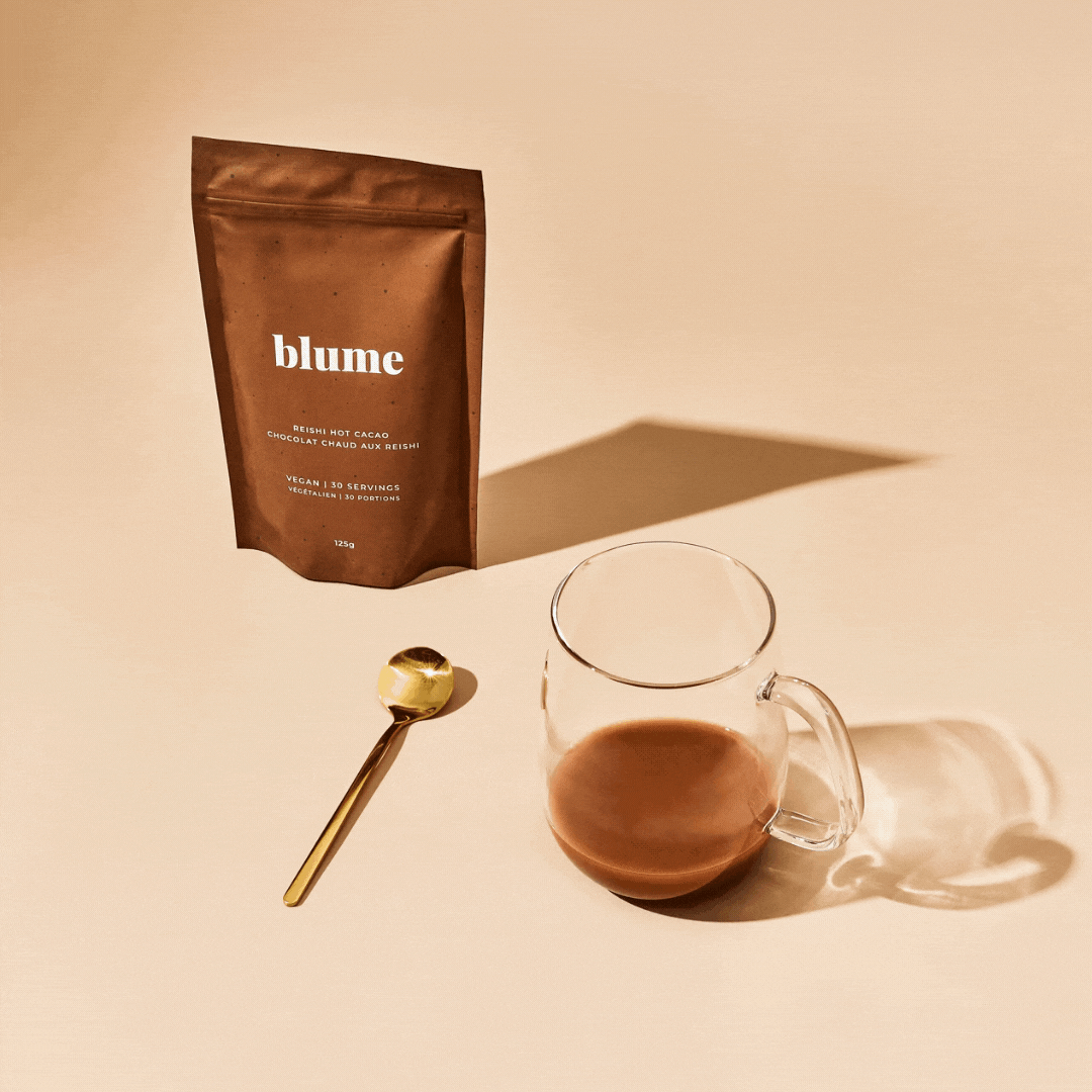 A gif of a glass mug filled up with hot chocolate, with a bag of Blume Reishi Hot Cacao Blend and a gold spoon on a beige background