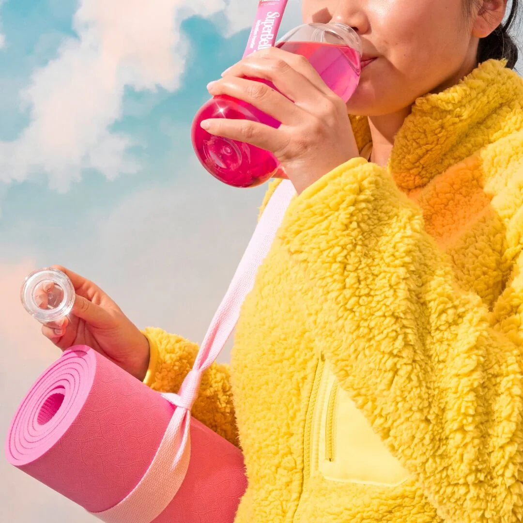 A person in a yellow fleece jacket drinks a pink beverage from a bottle marked Hydrate. They hold a rolled pink yoga mat with a white strap. The background shows a sky with clouds.