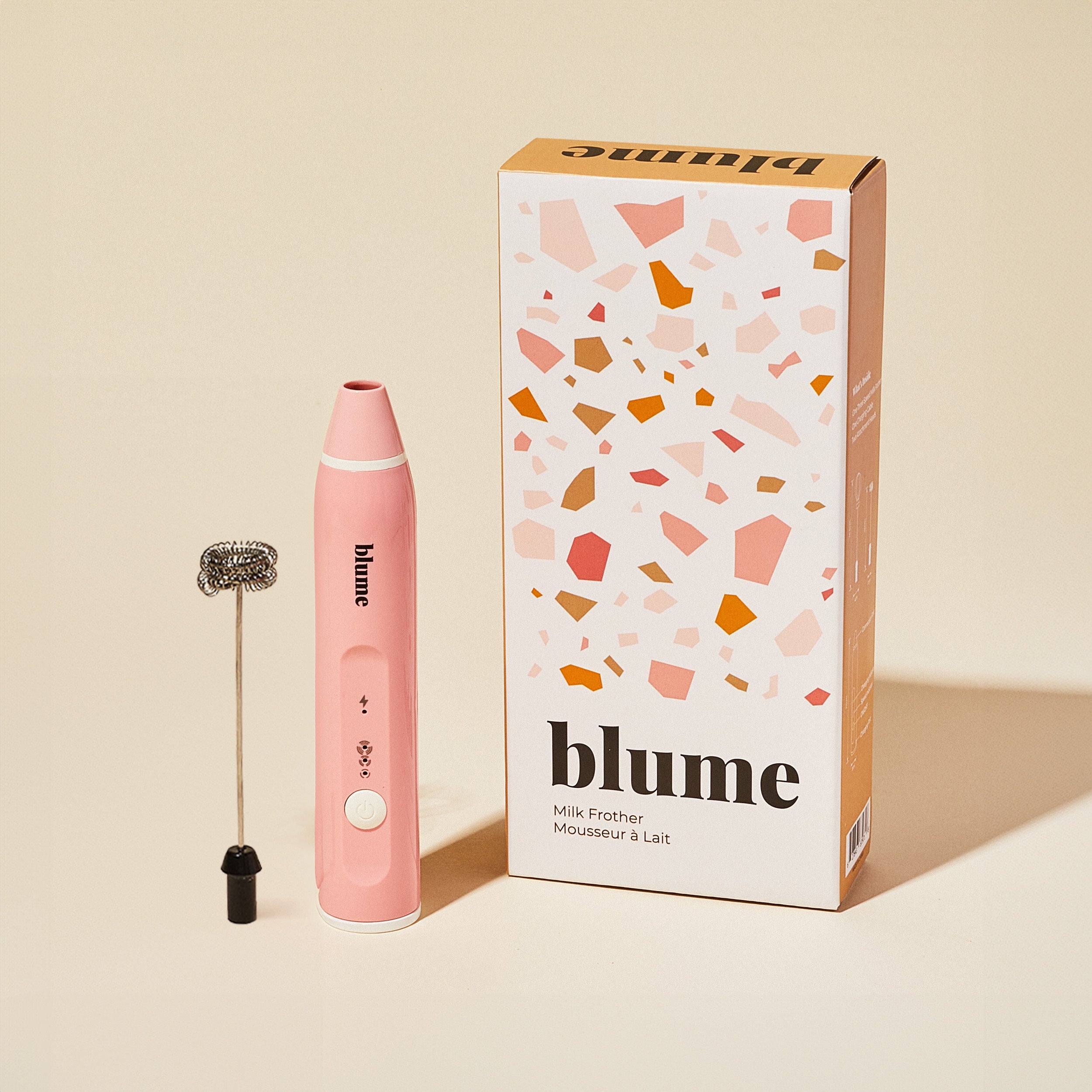 A pink Blume 3-Speed Milk Frother stands upright with its detachable metal whisk beside packaging. The device features USB charging, and the box showcases abstract pink and orange patterns with the brand name Blume in bold letters.
